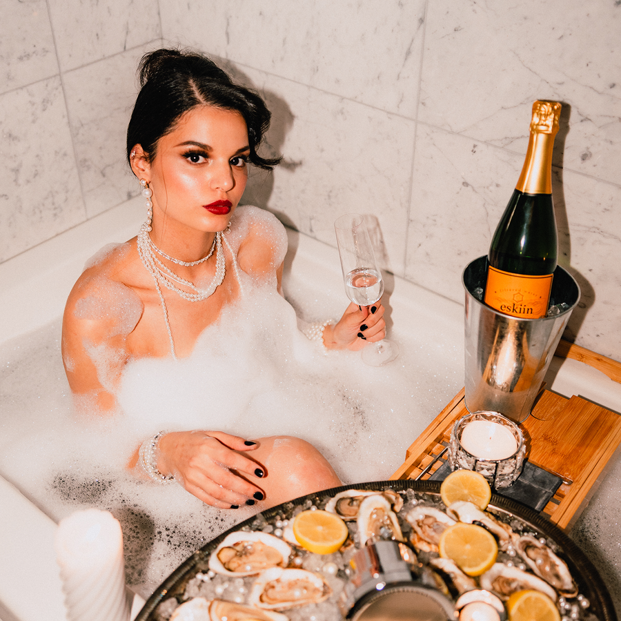 Woman in bathtub with champagne, pearls, and seafood platter.